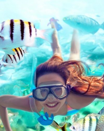 Smiling woman snorkeling underwater among colorful tropical fish in clear blue water, enjoying marine life in Cancun, Mexico.